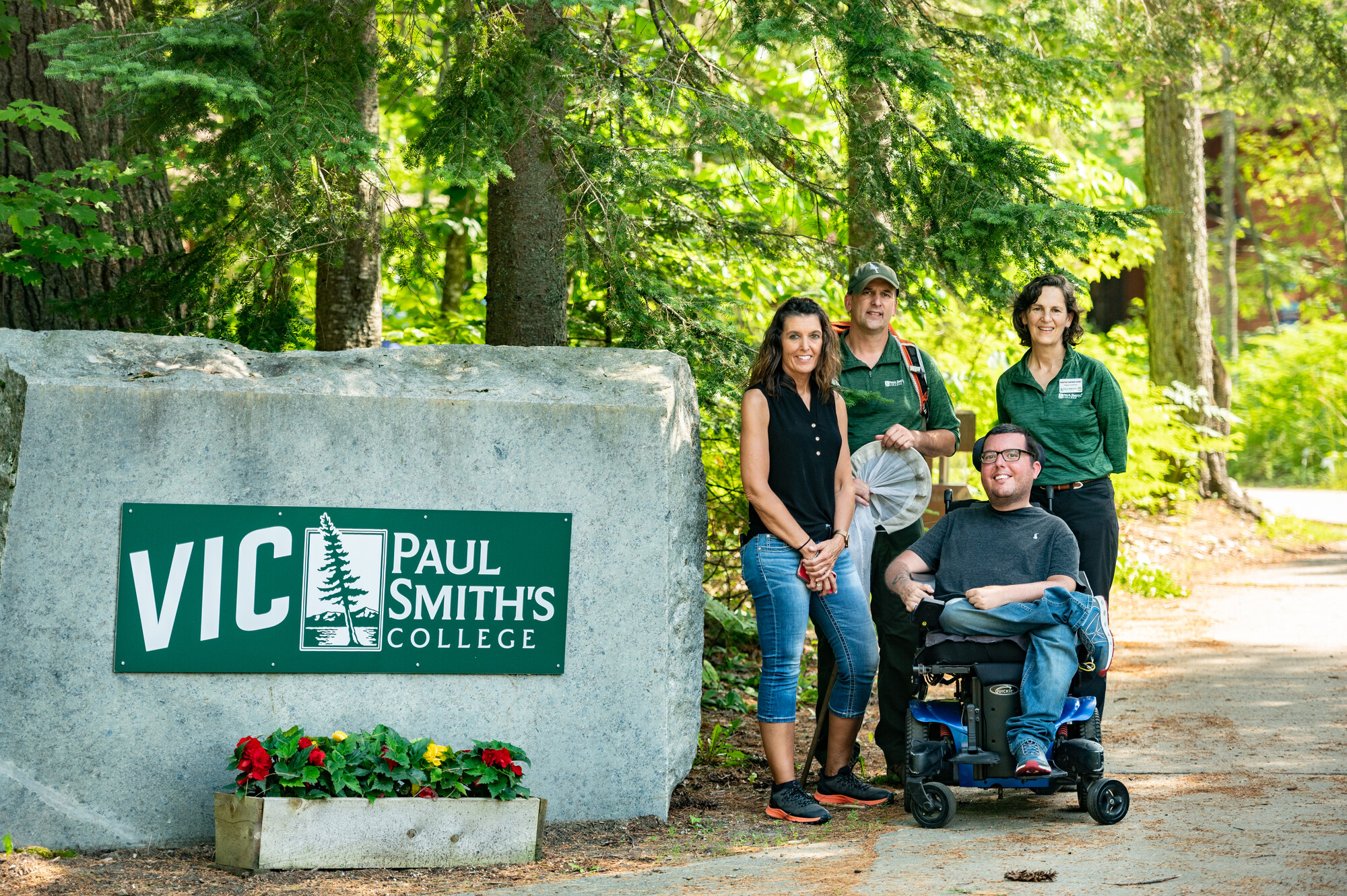 Travel Writer Corey Lee from Curb Free with Cory Lee poses with his mother and two staff members from Paul Smith's College VIC next to the entrance sign on a beautiful summer day..